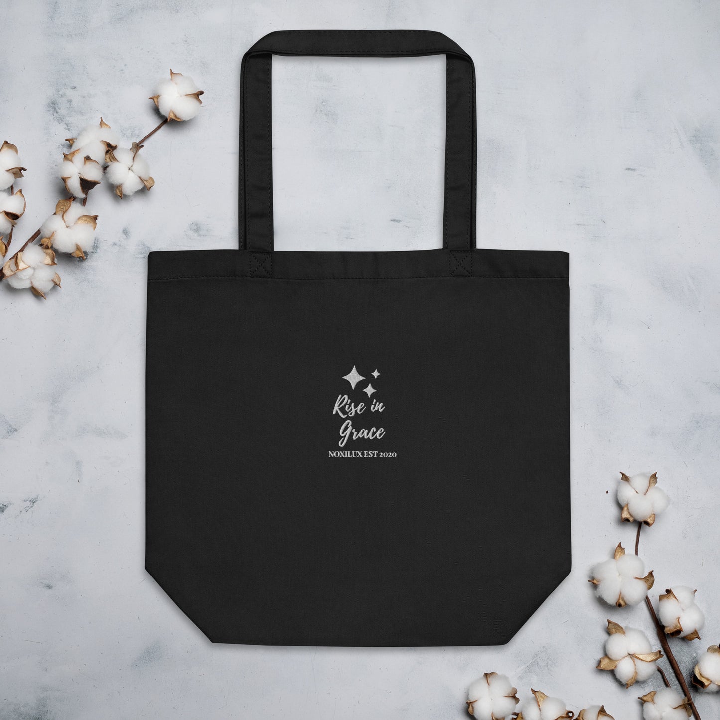 Black shopping tote bag with 'rise in grace.' text and a sparkle symbol on a white embroidered. Eco-friendly organic cotton.